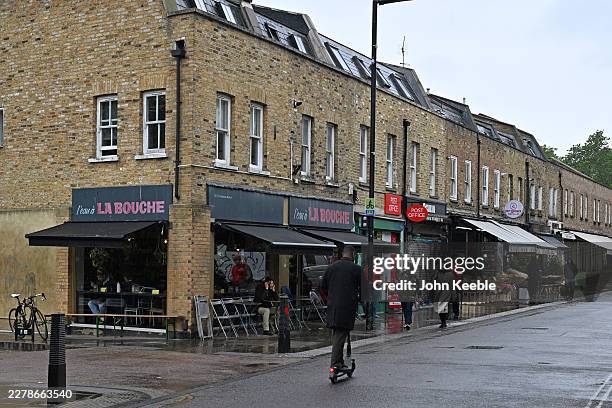 General view as people walk and a man scoots on a electric scooter on a wet day past shops and cafes along Broadway Market in the London Borough of...