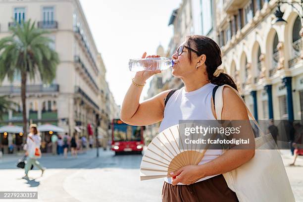 femme qui boit de l’eau près de la rue de la ville, souffrant de déshydratation et de coup de chaleur pendant les températures estivales élevées. vague de chaleur - éventail photos et images de collection