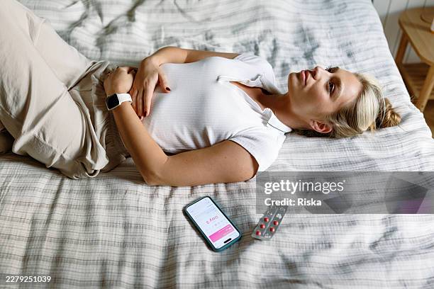 young woman resting on bed with period pain, phone app and medication for menstruation and woman's health - hormone stock pictures, royalty-free photos & images