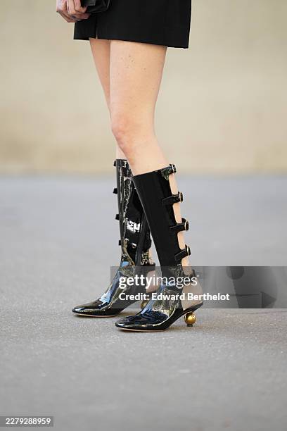 Close-up detail view of black shiny patent leather pointed-toe knee-high boots shoes with cutout sides, five slim strap details with round gold-tone...