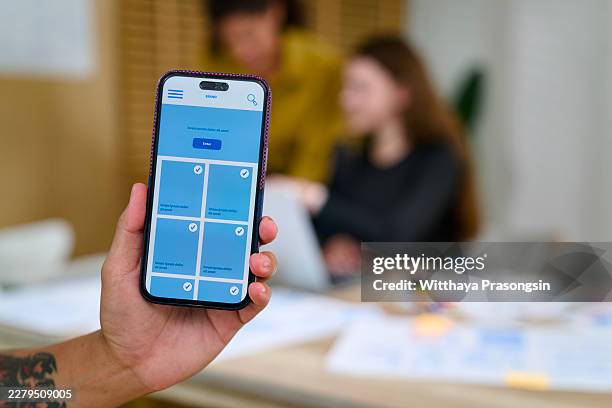 hand holding smartphone displaying mobile app prototype during team collaboration, developing digital product design with user interface and experience in office setting - prototipo fotografías e imágenes de stock