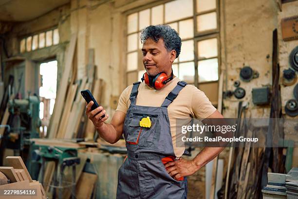 carpenter using smartphone in busy woodworking workshop - handyman stock pictures, royalty-free photos & images