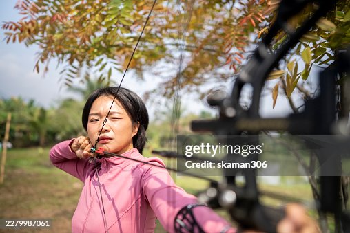 Focused young Asian woman draws a Compound Bow bow while practicing archery outdoors. Surrounded by green trees, she carefully aims the arrow, showing concentration, strength, and outdoor sporting activity. Focused young Asian woman draws a Compound Bow bow while practicing archery outdoors. Surrounded by green trees, she carefully aims the arrow, showing concentration, strength, and outdoor sporting activity.