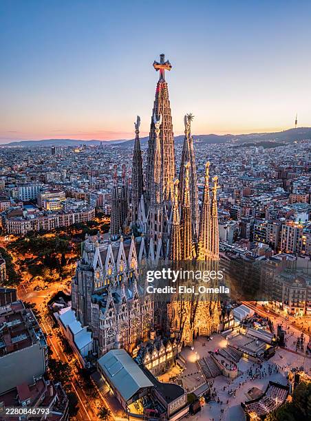sagrada familia basilica illuminated at sunset, standing tall against the dramatic twilight sky, with the sprawling barcelona cityscape visible below from an aerial perspective - sagrada família imagens e fotografias de stock