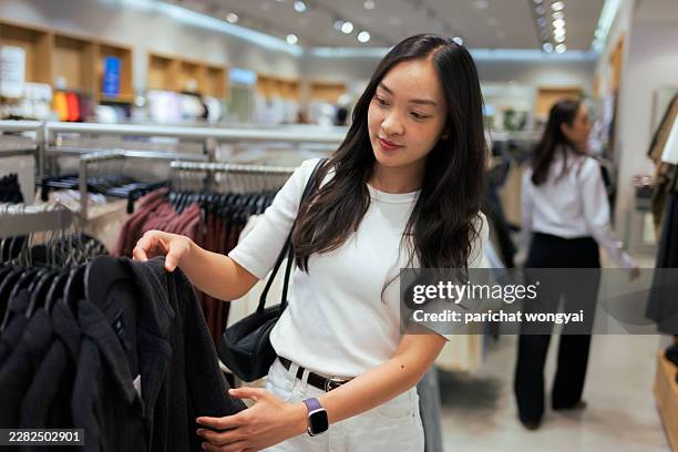 young adult woman checking black garment on a hanger in a brightly lit retail clothing store - shirt dress stock pictures, royalty-free photos & images