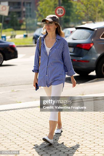 Guest wears a grey baseball hat, a white tank top, a light blue striped shirt, a yellow bag, white Capri pants and white ballerinas outside Pronounce...