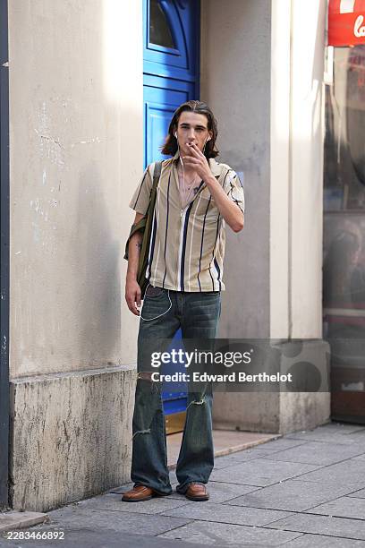 Guest wears long straight brown hair, a beige striped print pattern short-sleeve shirt, blue faded / ripped flared jeans, a green shoulder bag, brown...