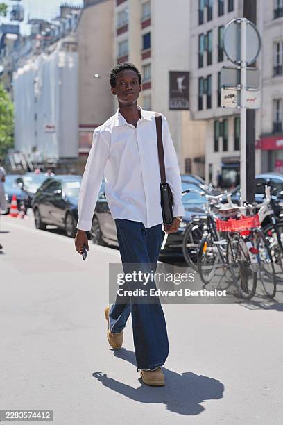 Guest wears short curly black hair, a white shirt, blue denim ripped wide-leg jeans, a black leather crossbody bag, tan suede boots shoes, outside...