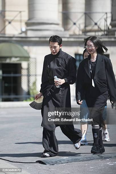 Guest wears short straight black hair, a black striped print pattern high collar shirt, matching black striped wide-leg pants, a black denim knees...