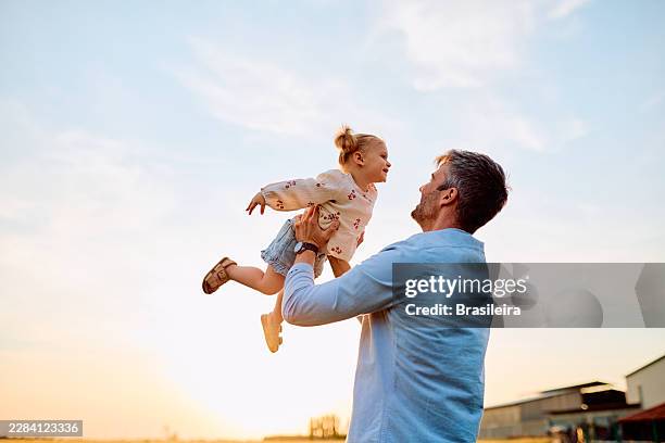 father lifting daughter playing outdoors during sunset - back lit stock pictures, royalty-free photos & images