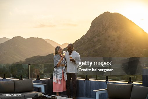 Totalbild eines erwachsenen Paares, das mit Wein auf der Dachterrasse bei Sonnenuntergang anstoßt Totalbild eines erwachsenen Paares, das mit Wein auf der Dachterrasse bei Sonnenuntergang anstoßt