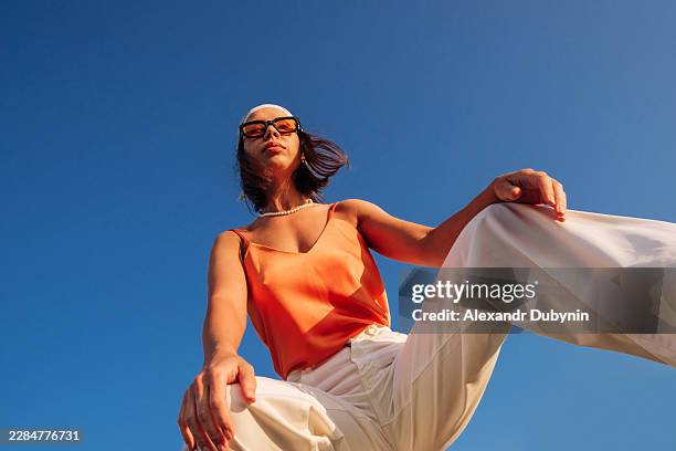 portrait of a fashionable woman squatting and looking at the camera against the blue sky in summer clothes - white pants stock pictures, royalty-free photos & images