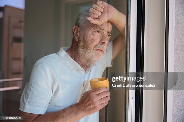 senior man feeling lonely looking through window - social isolation stock pictures, royalty-free photos & images