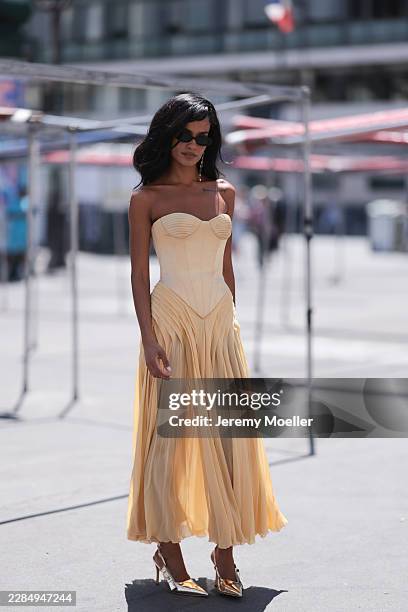 Narah Baptista is seen wearing narrow black oval sunglasses, a single long gold drop earring, a strapless butter-yellow corset dress with a sculpted...