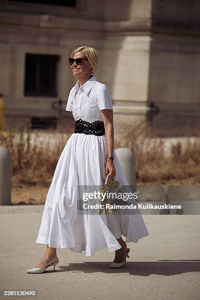 Guest wears a white short-sleeved shirt dress with a full skirt, a black floral cutout waist belt, burgundy sunglasses, carries a beige top-handle...