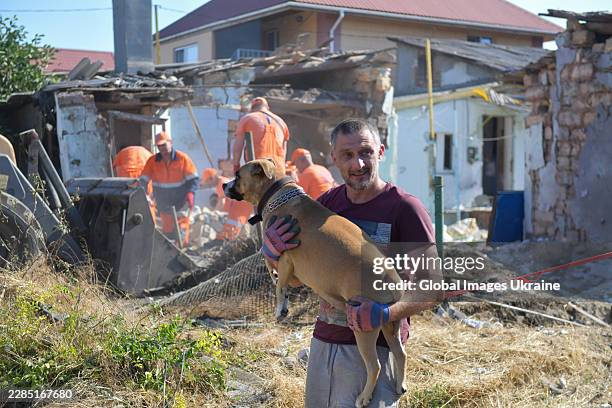 Man holds a dog in his arms in front of workers clearing rubble from a house partially destroyed by a Russian strike on July 6, 2026 in Odesa,...