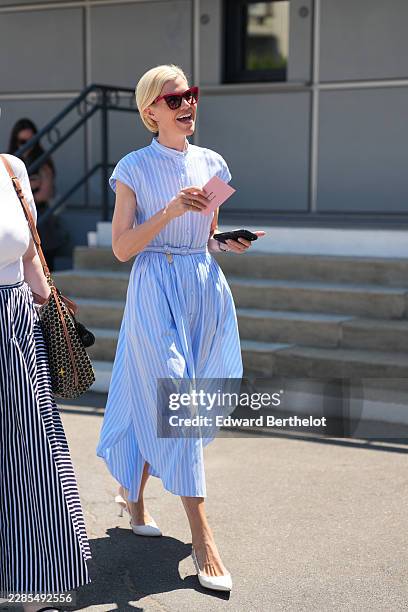 Guest wears short blonde bob hair, red cat-eye sunglasses, a blue striped print pattern short sleeves / belted midi shirt dress with a belt, white...