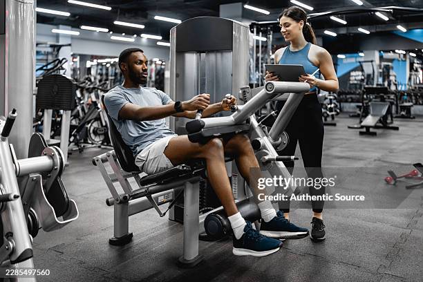 man training with personal trainer at gym using leg machine while coach records workout - hamstring machine stock pictures, royalty-free photos & images