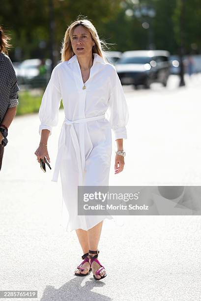 Guest is seen wearing a white belted midi shirt dress with voluminous sleeves; pink and black strappy flat sandals; delicate gold jewelry; holding a...