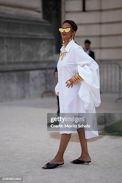 Guest is seen wearing a white oversized shirt dress; black pointed toe mules with gold kitten heels; mirrored gold cat eye sunglasses; a full...
