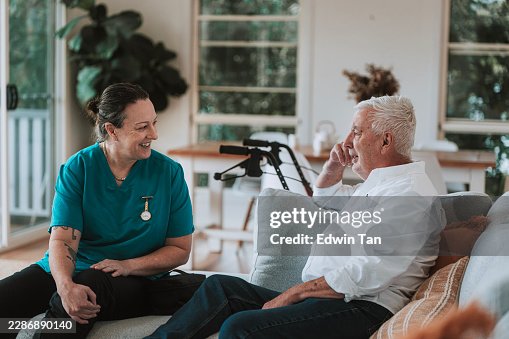 Häusliche Pflegekraft lacht mit älterem männlichen Patienten auf dem Sofa im Wohnzimmer während des Hausbesuchs Häusliche Pflegekraft lacht mit älterem männlichen Patienten auf dem Sofa im Wohnzimmer während des Hausbesuchs