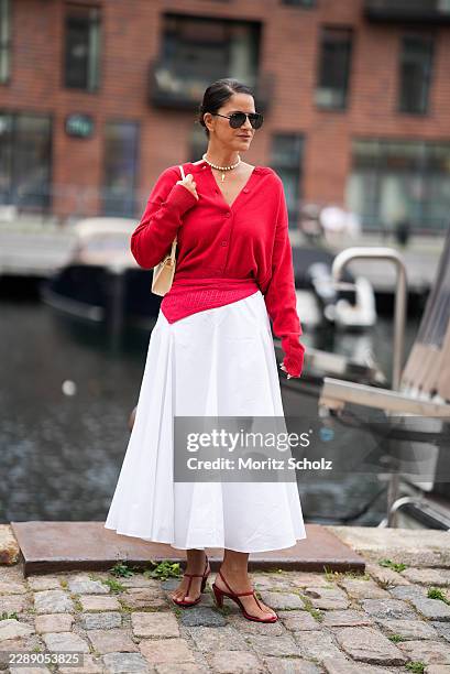 Gili Biegun is seen wearing a red knit cardigan with a V neck; a white full midi skirt; red leather thong sandals with high heels; a small cream...