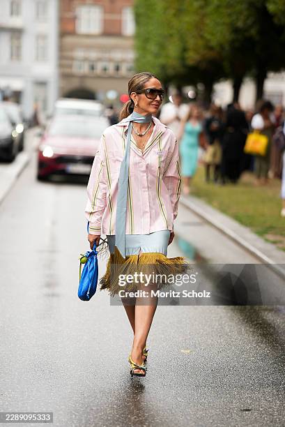 Tiany Kiriloff is seen wearing a light pink striped button up shirt; a light blue silk scarf tied around the neck; a light blue skirt with an olive...
