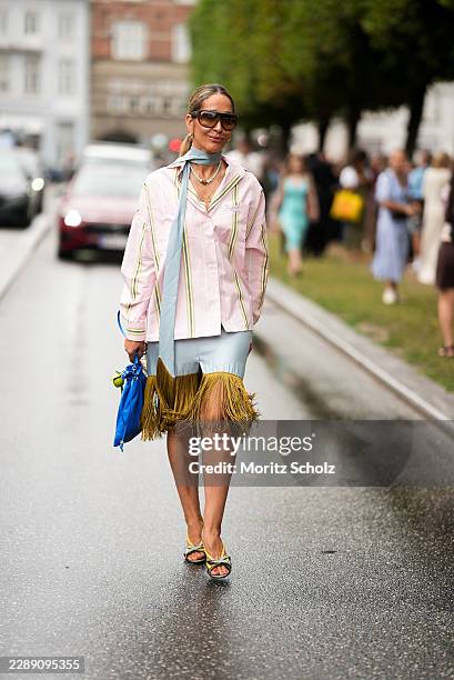 Tiany Kiriloff is seen wearing a light pink striped button up shirt; a light blue silk scarf tied around the neck; a light blue skirt with an olive...
