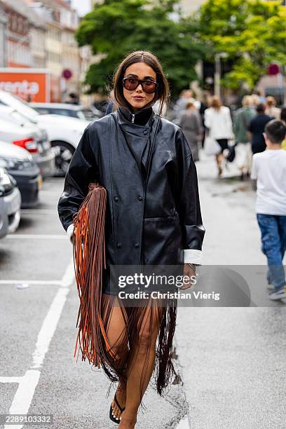 Karen Gonzalez wears black leather coat, fringed brown jacket, dress, sunglasses outside MKDT Studio during the Copenhagen Fashion Week Spring/Summer...