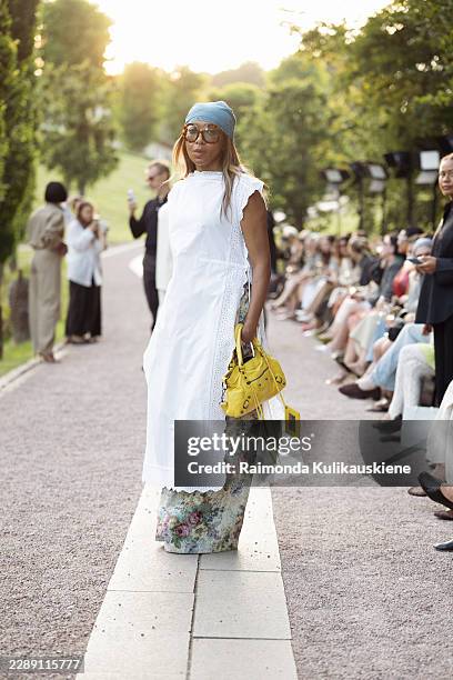 Karen Blanchard wears a turquoise bandana with oversized sunglasses, a white eyelet wrap dress layered over floral wide leg trousers, a bright yellow...