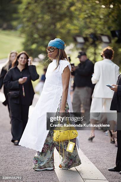 Karen Blanchard wears a turquoise bandana with oversized sunglasses, a white eyelet wrap dress layered over floral wide leg trousers, a bright yellow...