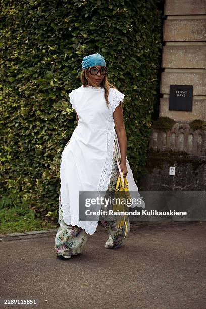 Karen Blanchard wears a turquoise bandana with oversized sunglasses, a white eyelet wrap dress layered over floral wide leg trousers, a bright yellow...
