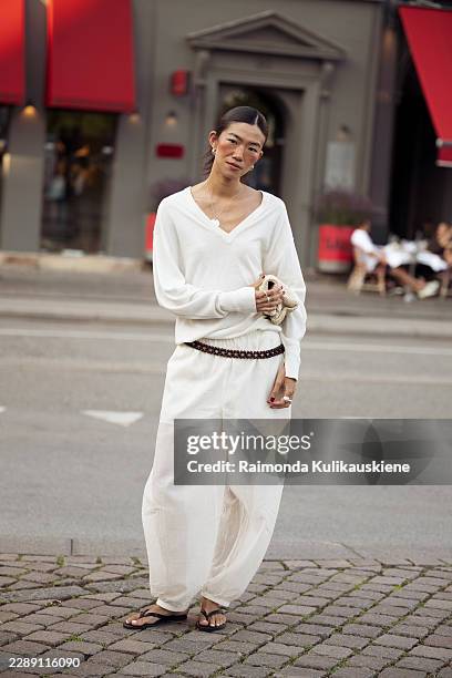 Guest wears an ivory V neck sweater with matching wide leg trousers, a dark beaded belt, silver jewelry, a beige knitted clutch and black flip flop...