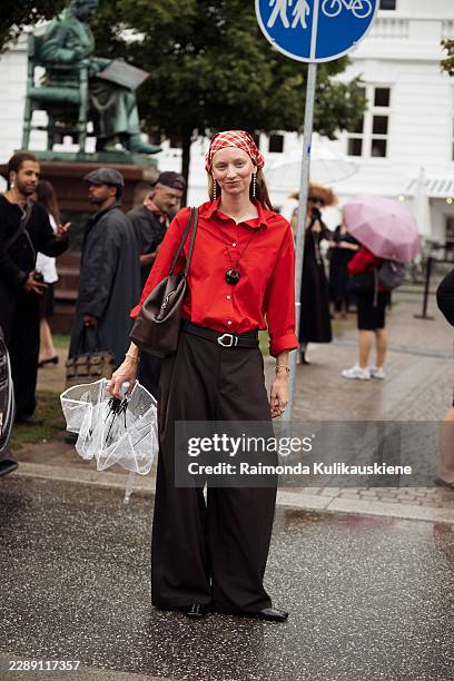 Laura Schulte wears wide brown pants, red shirt, brown belt, red and white head scarf, black round necklace, transparent umbrella and brown bag...