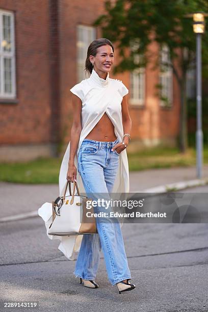 Guest wears long straight light brown hair with a side part, a white satin draped turtleneck sleeveless cropped top with a long back panel cape,...