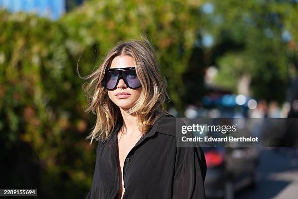 Guest wears shoulder-length straight blonde hair with a center part, oversized black shield sunglasses with blue colored lenses, a black long-sleeve...