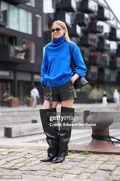 Guest wears long straight blonde hair with a center part, black rectangular sunglasses with dark lenses, a cobalt blue knit sweater with oversize...