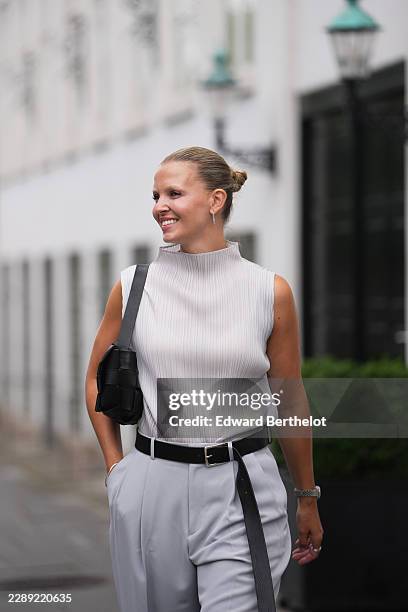 Hollie Mercedes Peters wears blonde slicked-back hair in a bun, a sleeveless light gray pleated mock-neck top, light gray wide-leg pants, a black...