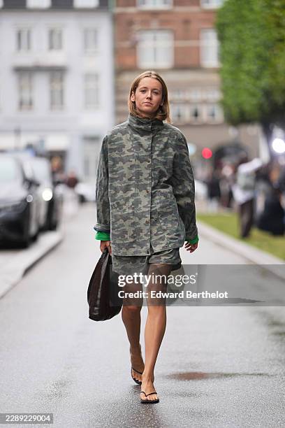 Guest wears blonde shoulder-length straight hair with a center part, a green and gray camouflage print high-neck button-front jacket, over a green...