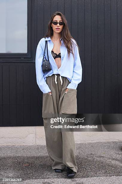 Renee Marianna van Bommel wears long wavy brown hair with a side part, black oval sunglasses with dark lenses, a light blue cotton button-through...