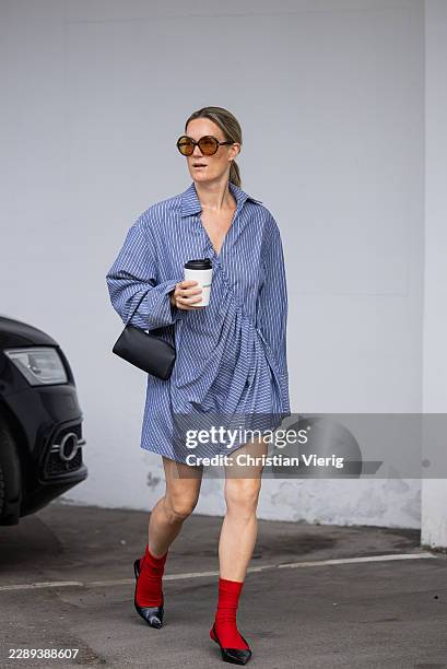 Victoria Thomas with coffee cup wears oversized blue striped button shirt dress, black bag, big orange toned sunglasses, red socks, pointed heels...