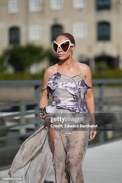 Guest wears brown hair in a slicked back bun, oversized white shield sunglasses with dark brown lenses, a strapless lilac silk and mesh corset top...