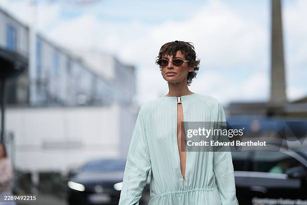 Irina Kro Eicke wears short wavy brown hair, oval silver-tone sunglasses with brown tinted lenses, a light blue long-sleeve midi dress with a deep...