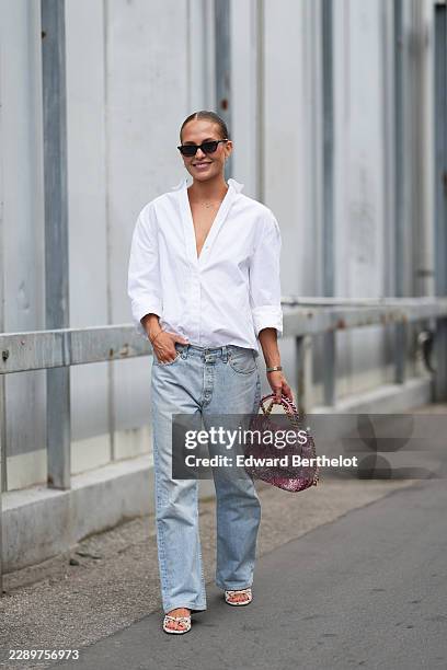 Guest wears blond slicked back hair in a bun, black rectangular sunglasses with dark lenses, a white cotton button-through shirt with an open collar...