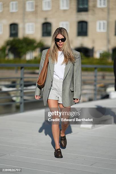 Guest wears long straight blonde hair with a center part, black cat-eye sunglasses with dark lenses, a gray and black houndstooth blazer jacket, a...