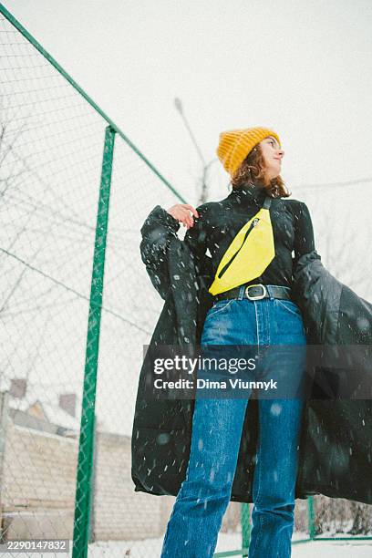 young woman in yellow beanie and bright sling bag standing outdoors during snowfall beside a chain link fence - sling bag stock pictures, royalty-free photos & images
