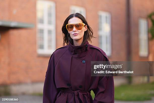 Guest wears shoulder-length wavy brown hair with a center part, amber shield sunglasses with tinted lenses, a burgundy long trench coat with a high...