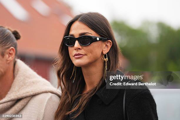 Guest wears long wavy dark brown hair with a center part, black oval sunglasses with dark lenses, a black wool coat, golden earrings, outside Skall...