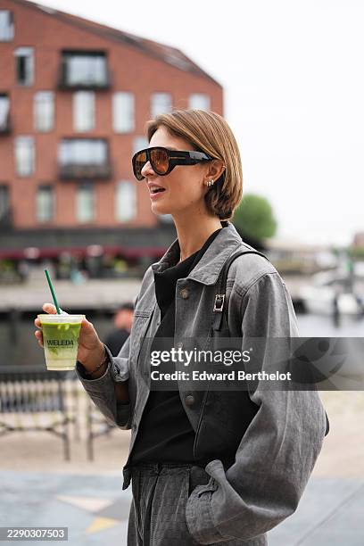 Guest wears short straight light brown bobbed hair with a center part, oversized black aviator sunglasses with tinted lenses, a charcoal gray denim...