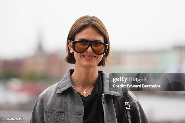 Guest wears short straight light brown bobbed hair with a center part, oversized black aviator sunglasses with tinted lenses, a charcoal gray denim...